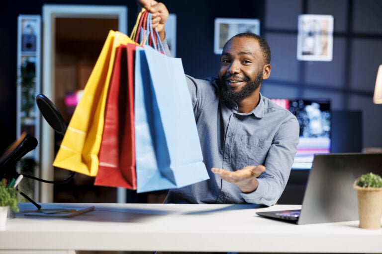 African american man filming fashion vlog, holding shopping bags in hand, showing subscribers purchases he recently got. Internet show host presenting viewers new acquisitions, filming himself at home